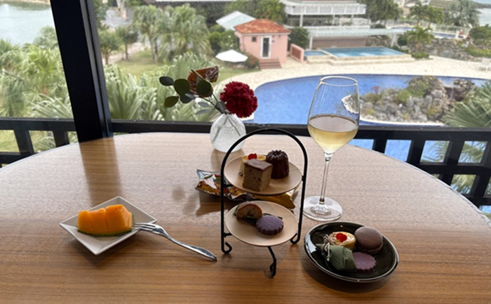 シギラミラージュのラウンジで提供されるミニアフタヌーンスイーツセット。窓外に広がるプールと海を背景にしたケーキスタンド、メロン、白ワイン